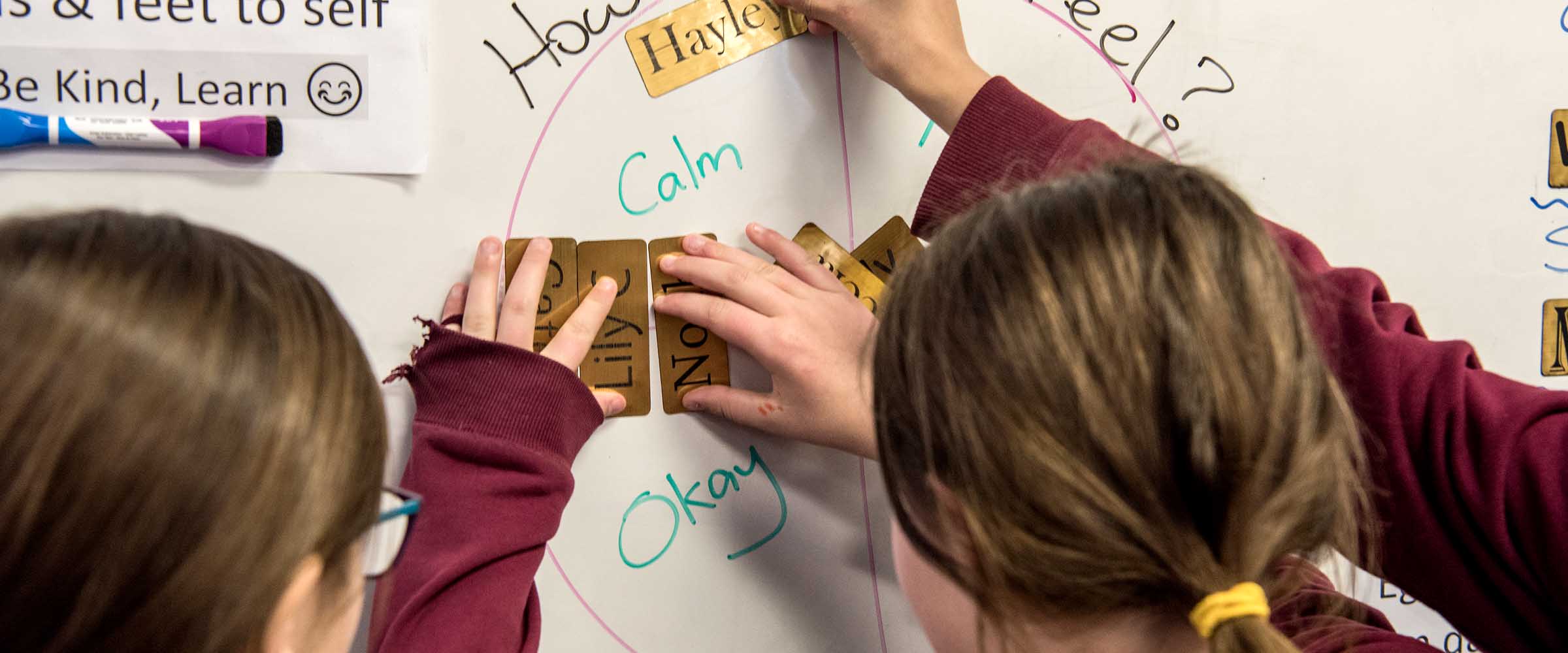 Three children standing in front of a whiteboard that has the words 'How do you feel?' written on it. The children are placing their names inside a circle on the whiteboard that says 'Calm' and 'OK'