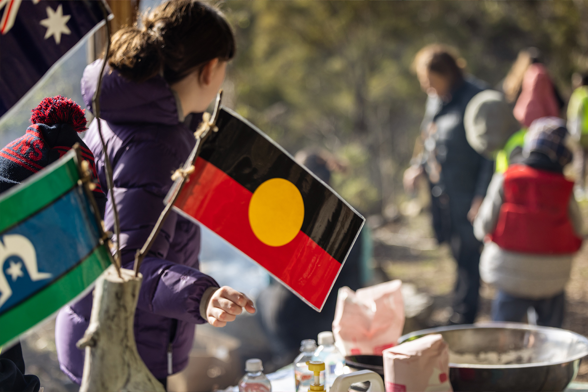 Image of a learning community outside going about activities in the background with the Aboriginal and Torres Strait Flags positioned in the foreground.