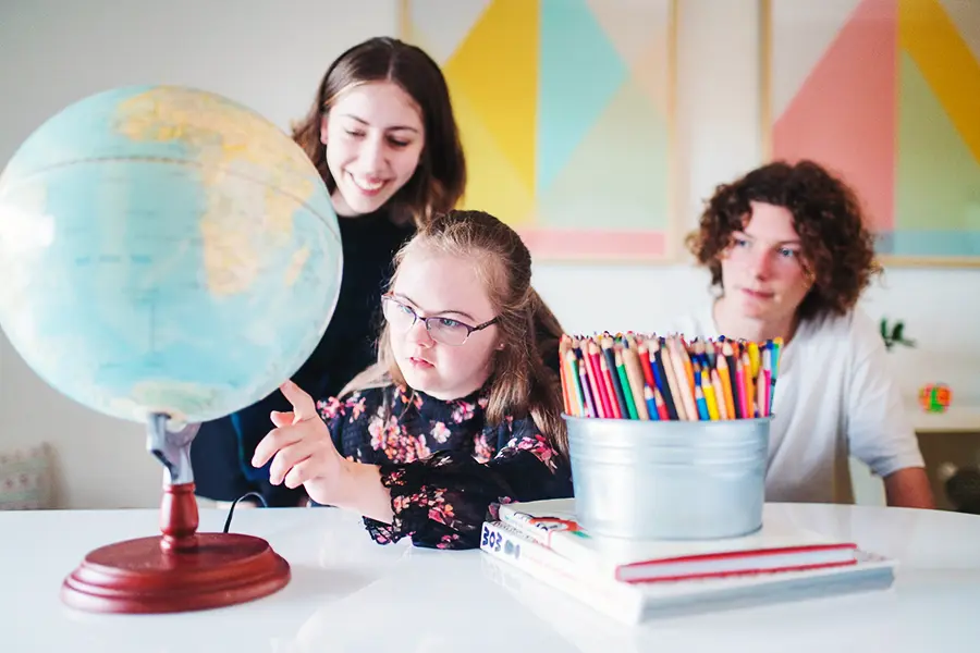 Three children sitting at a table with a globe, coloured pencils and books, representing inclusive learning, creativity and global curiosity in an early childhood setting.