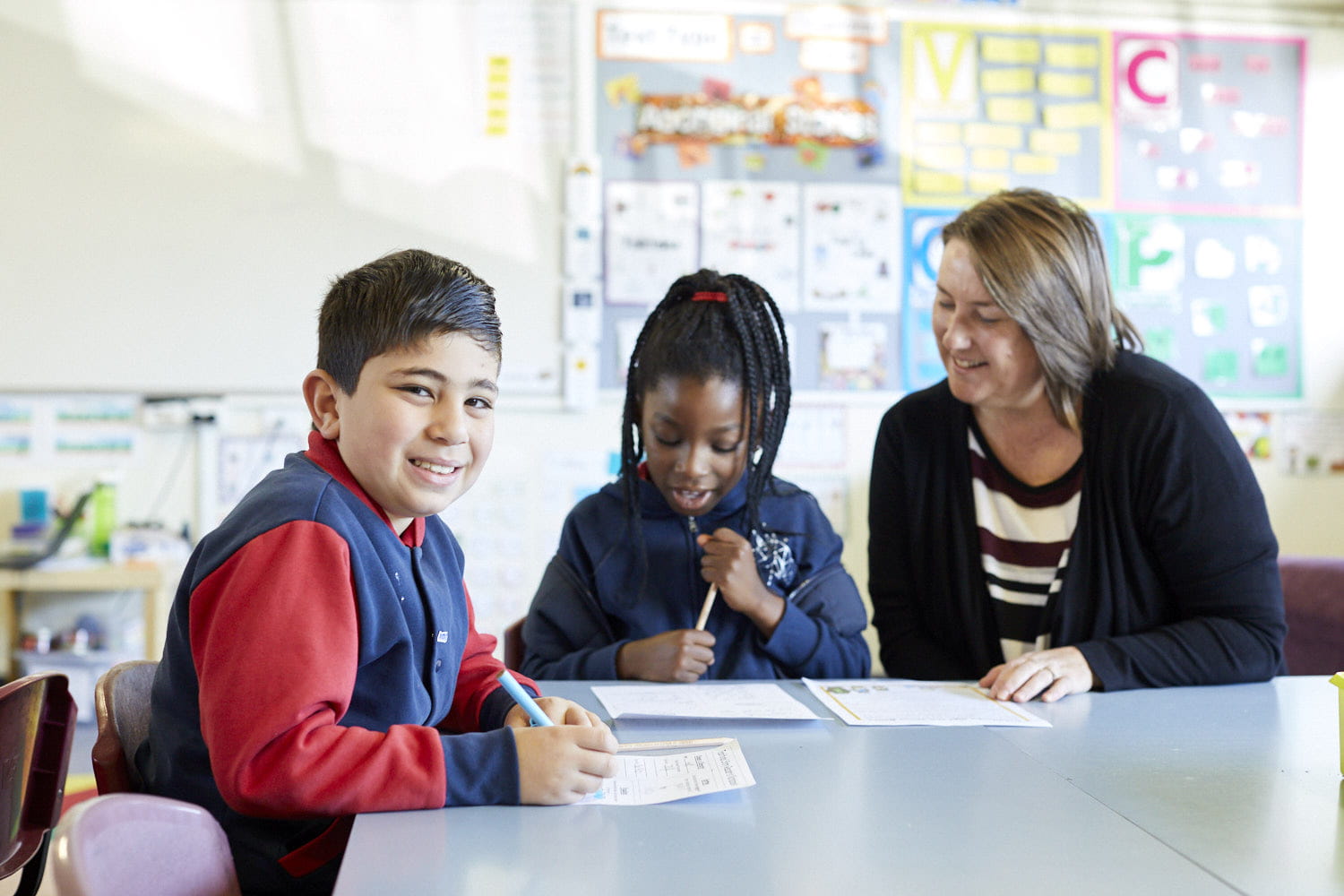 Two primary school aged students sitting at a tabke working with an adult.  One is looking at the camera and smiling.