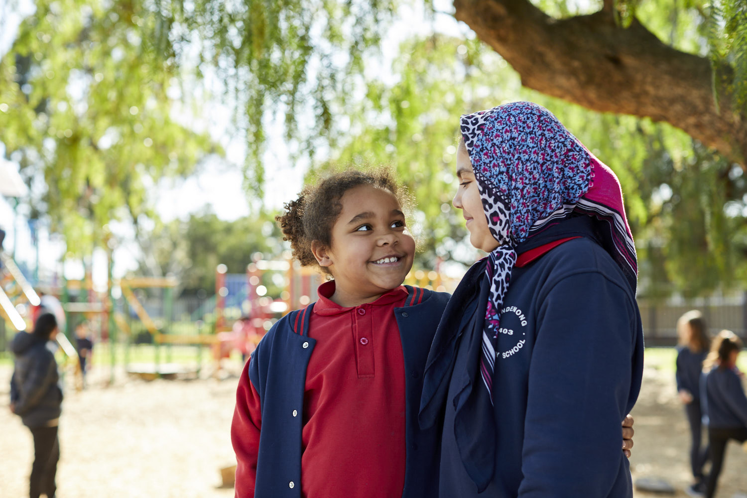 Two primary school aged students standing with their arms around each other in the playground, looking at each other and smiling.