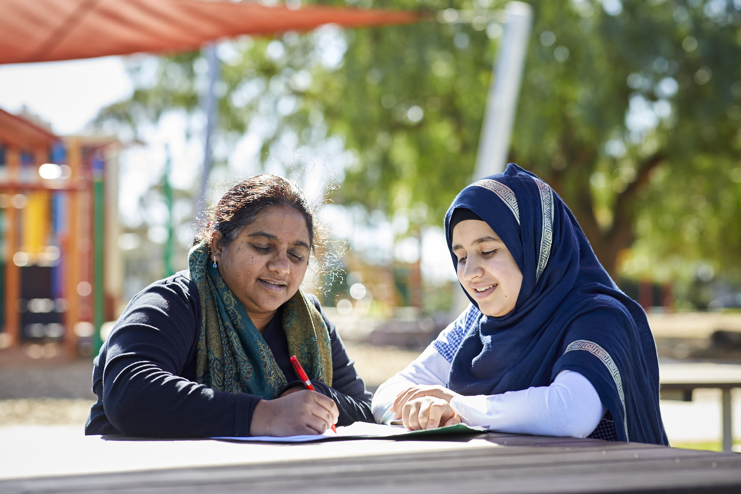 An educator and a student sit at a table outdoors with a playground in the background.