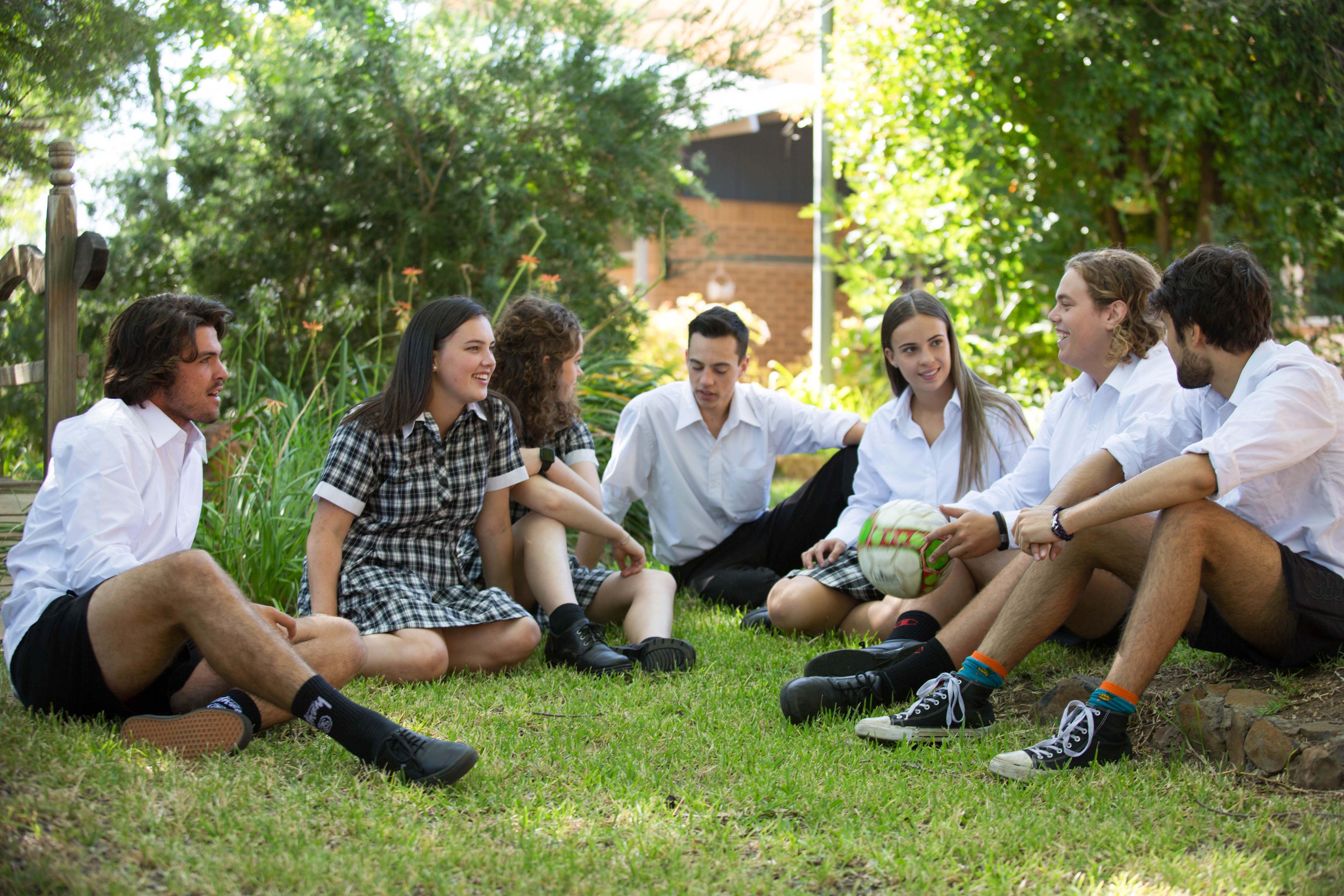 Seven teenage students sitting in a semi circle on the grass having a conversation.