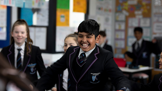 A primary school student in a classroom laughing with their arms spread out. There are smiling students in the background.