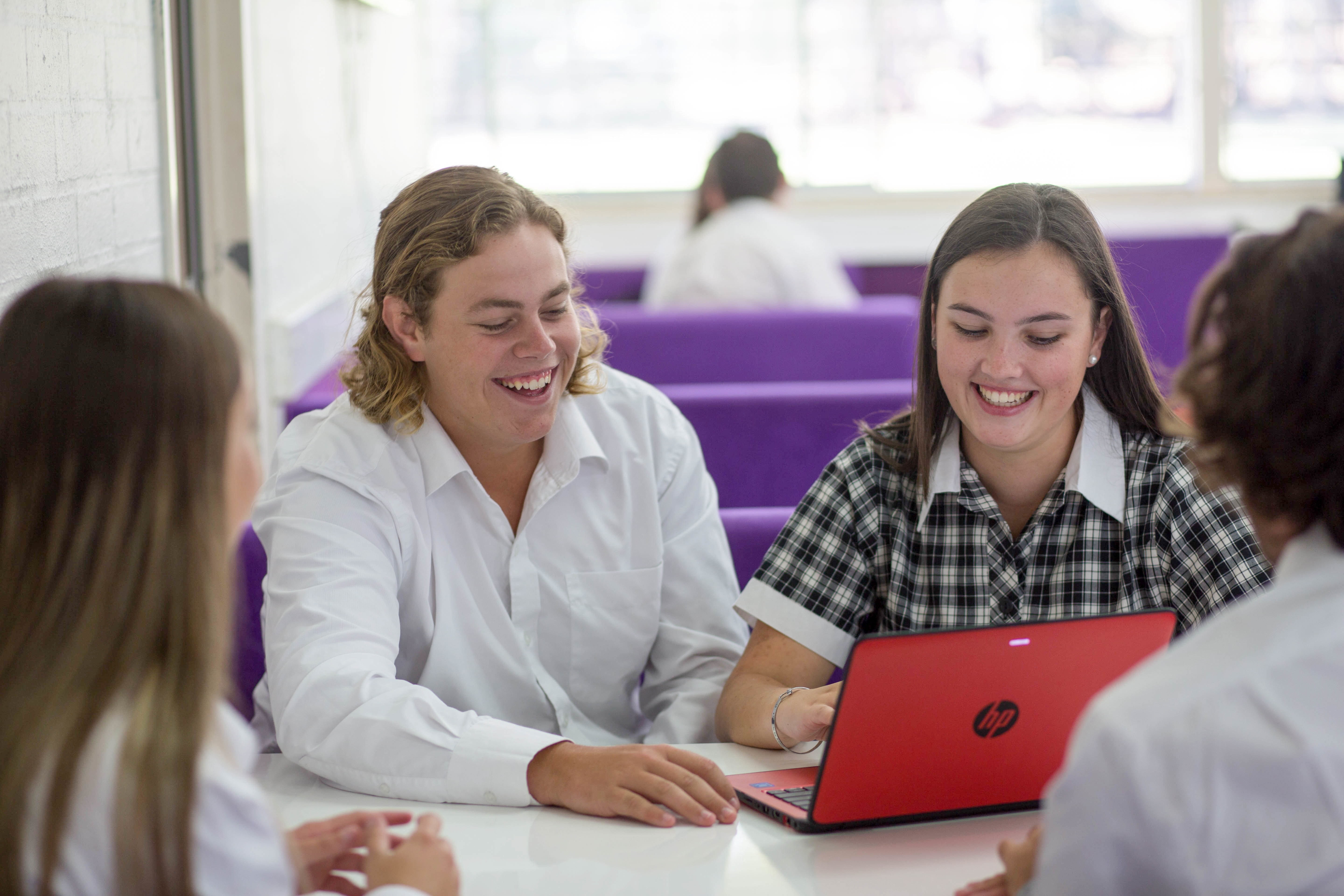 Four secondary aged students sitting in a booth together. Two are facing the camera and looking at a laptop together.