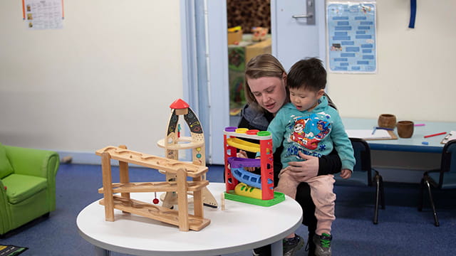 An adult sitting at a table covered in toys with a small child sitting on their lap.