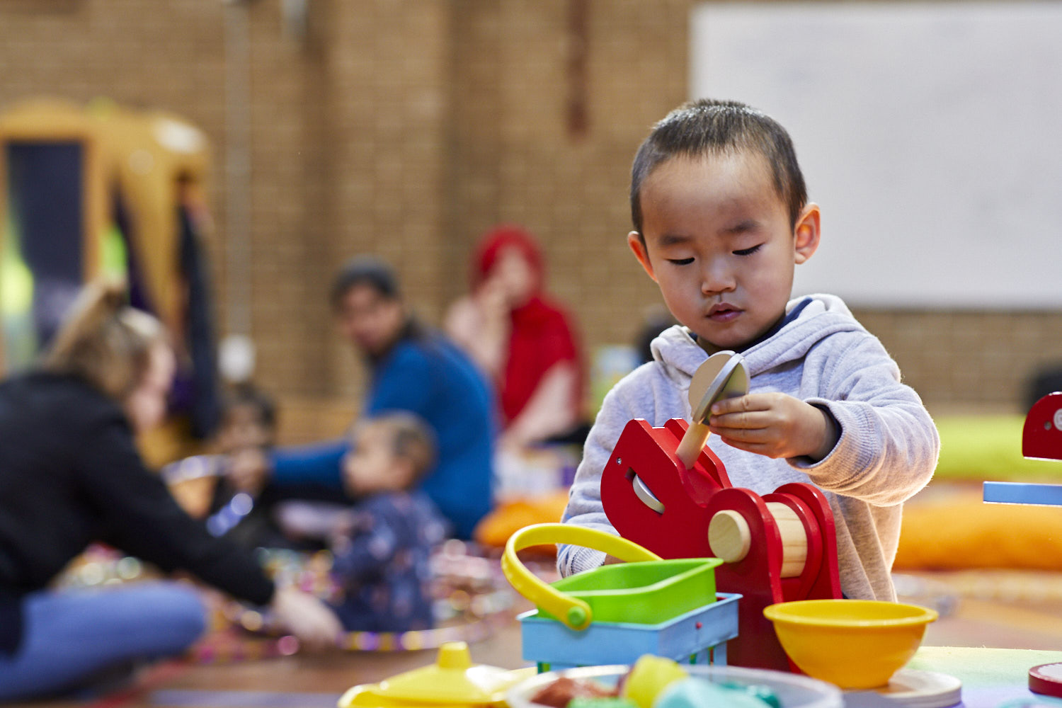 A young child sitting at a table with wooden toys. Adults and children are playing in the background.
