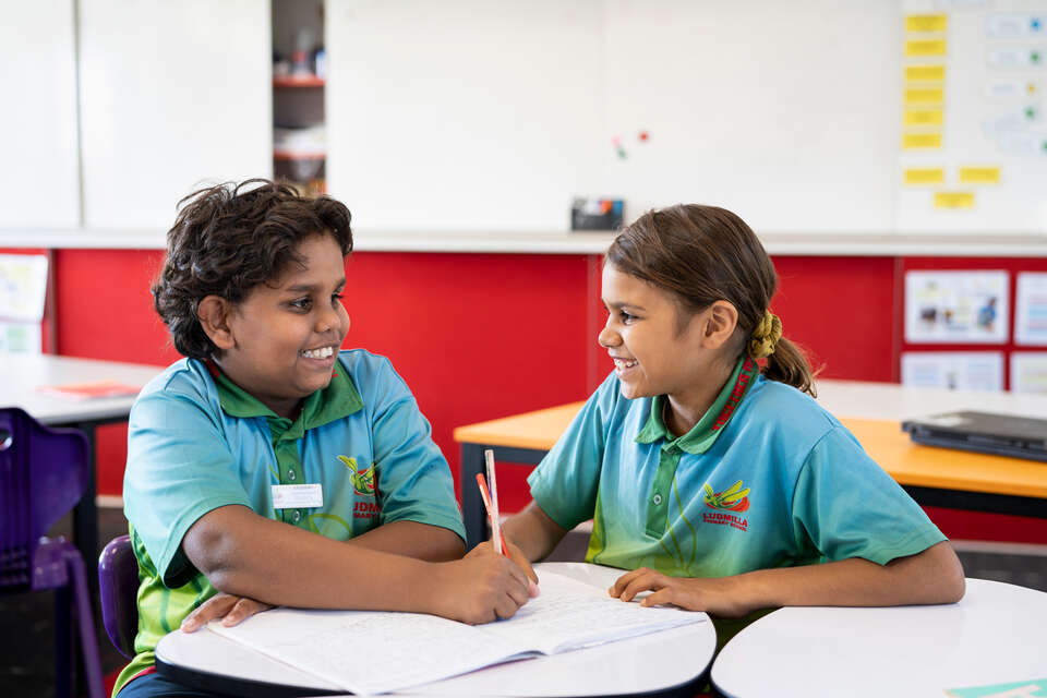 Two children, working at a desk, smiling at each other 