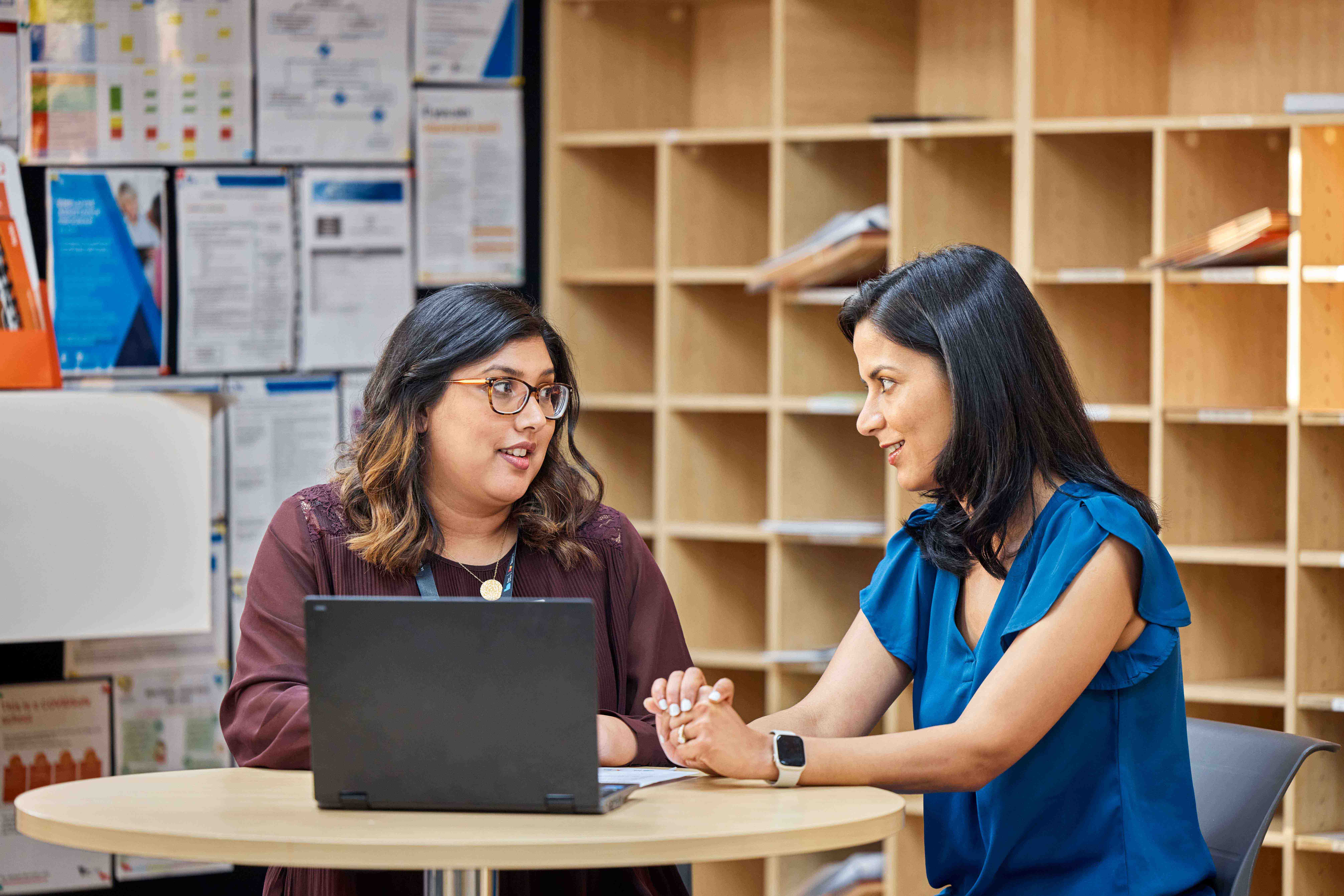 Two adults discussing at a table with a laptop in an office-like environment.