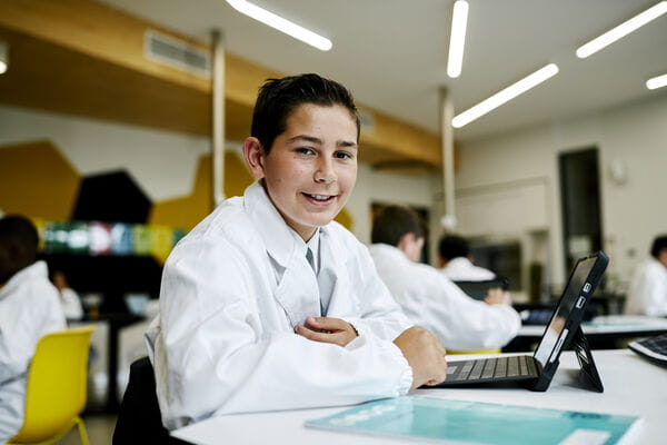 A smiling secondary school student working on their laptop in a school computer lab.