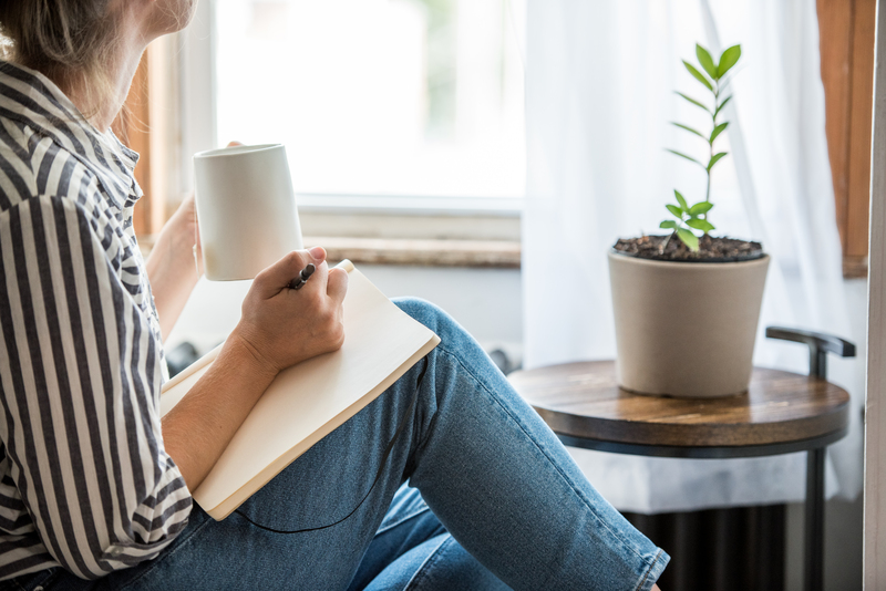 Woman sitting by a window writing in a journal and holding a mug.
