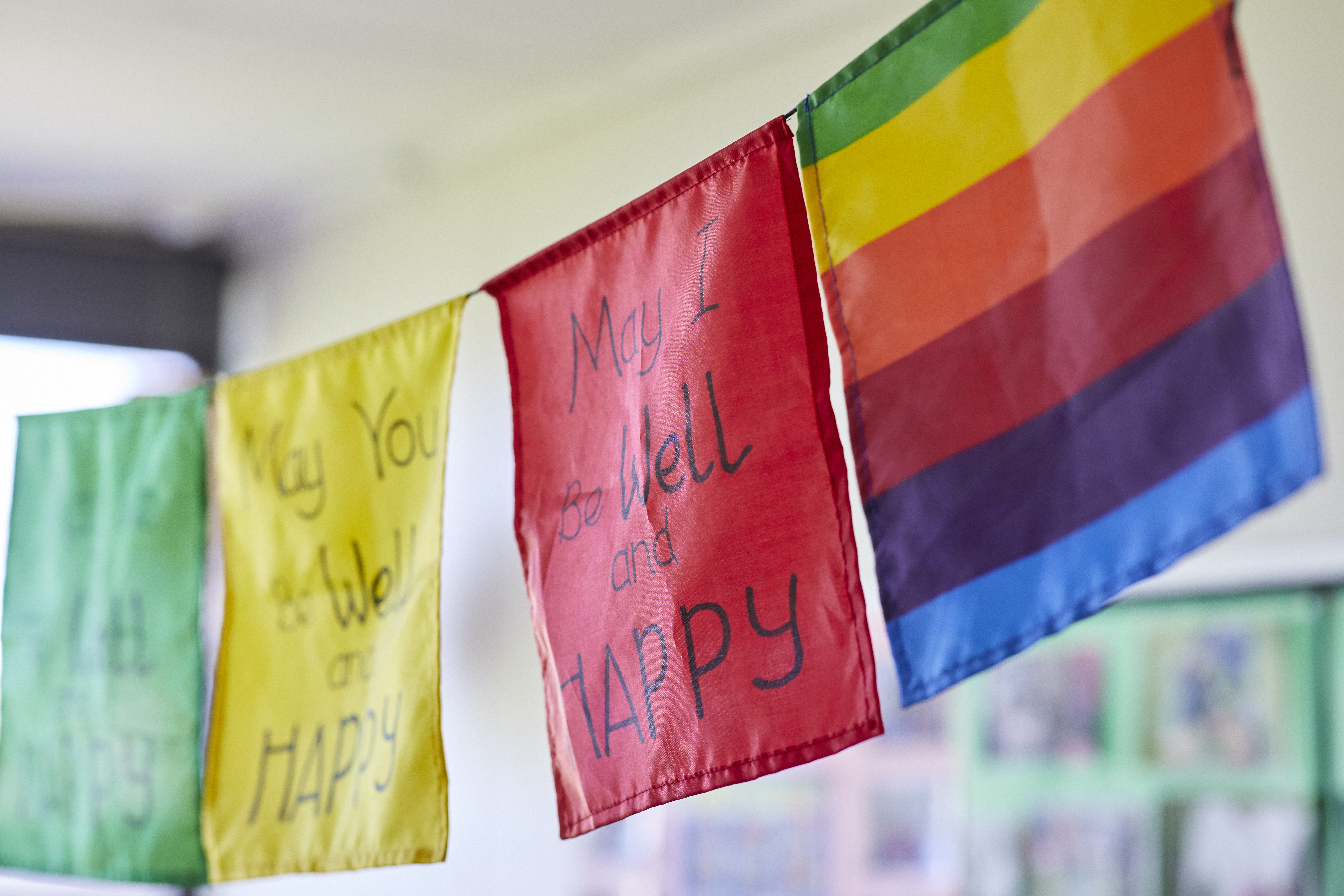 A green, yellow, red and rainbow flag hanging on a line, with may I be well and happy written on the red flag