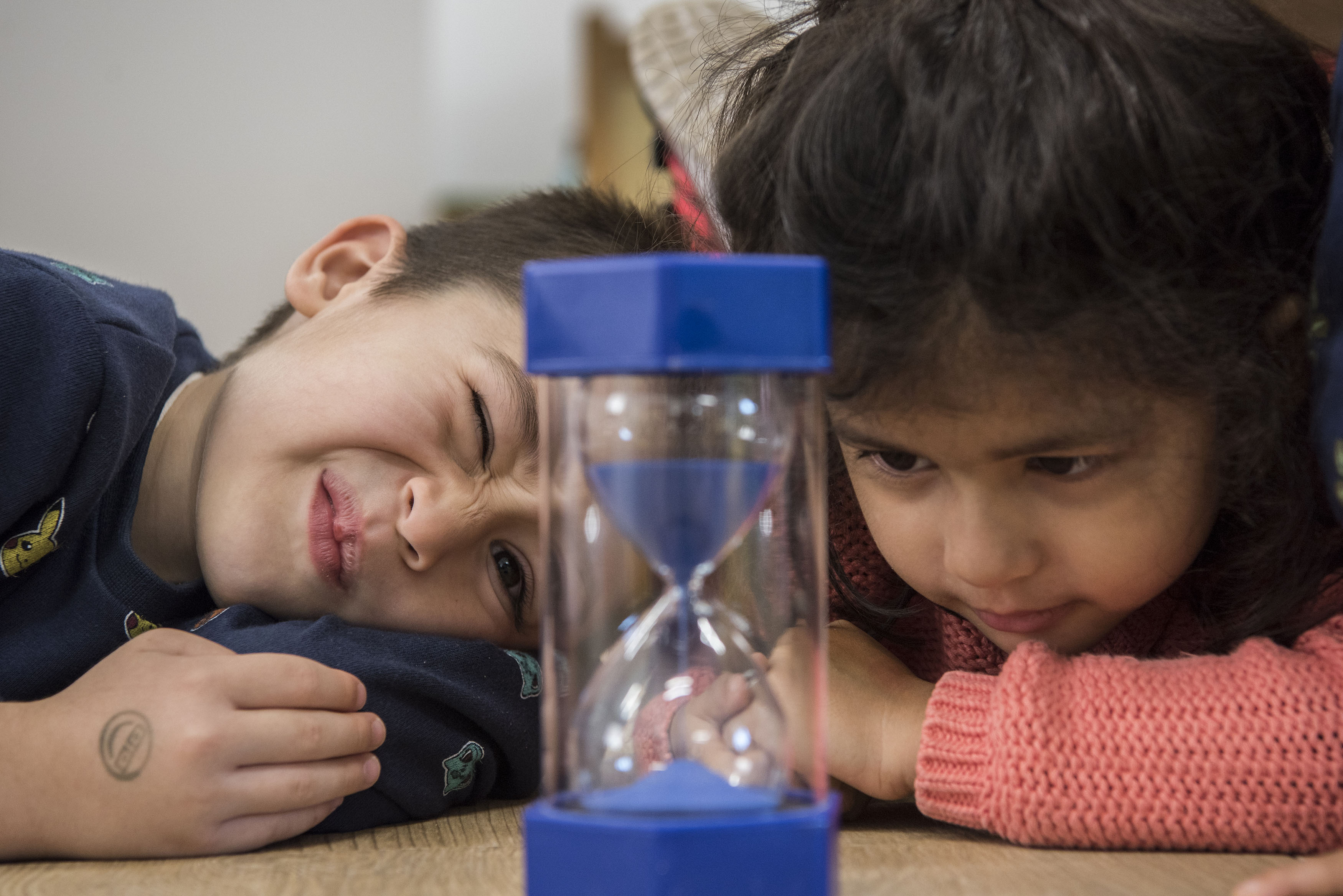 Two young children leaning on a table, looking closely at an hourglass