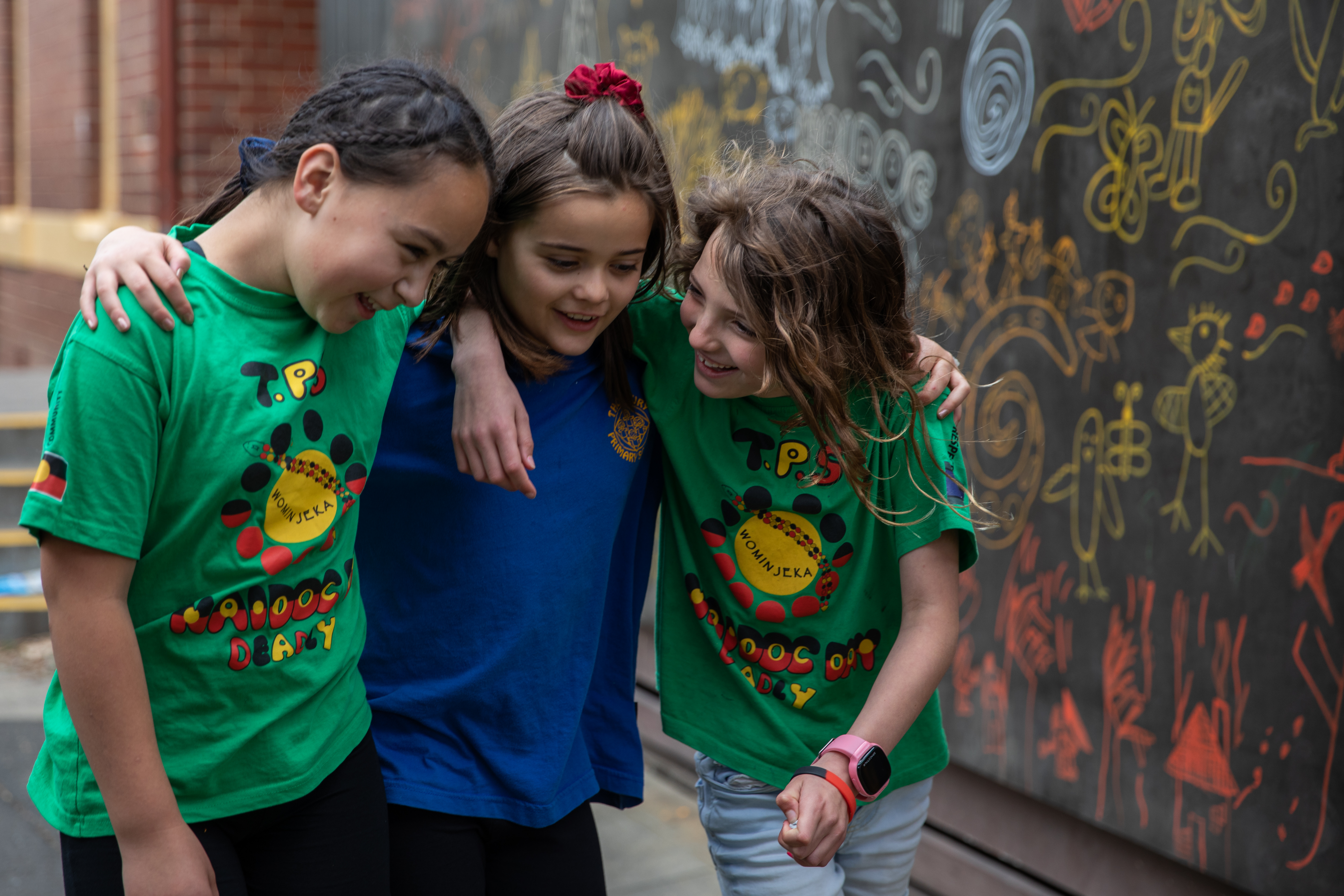 Three primary school aged students walking together with their arms around each other's shoulders. A colourful chalk art wall is in the background.