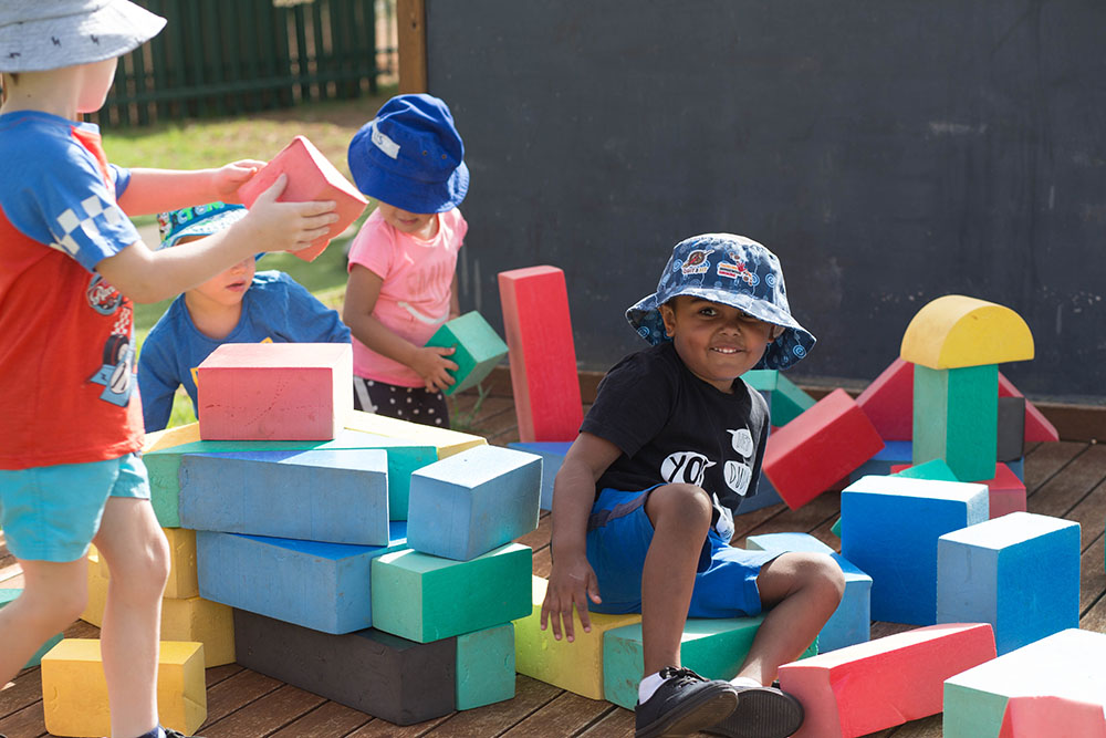 A group of four children playing together outside with colourful foam building blocks. One child is sitting on the blocks, looking at the camera and smiling.