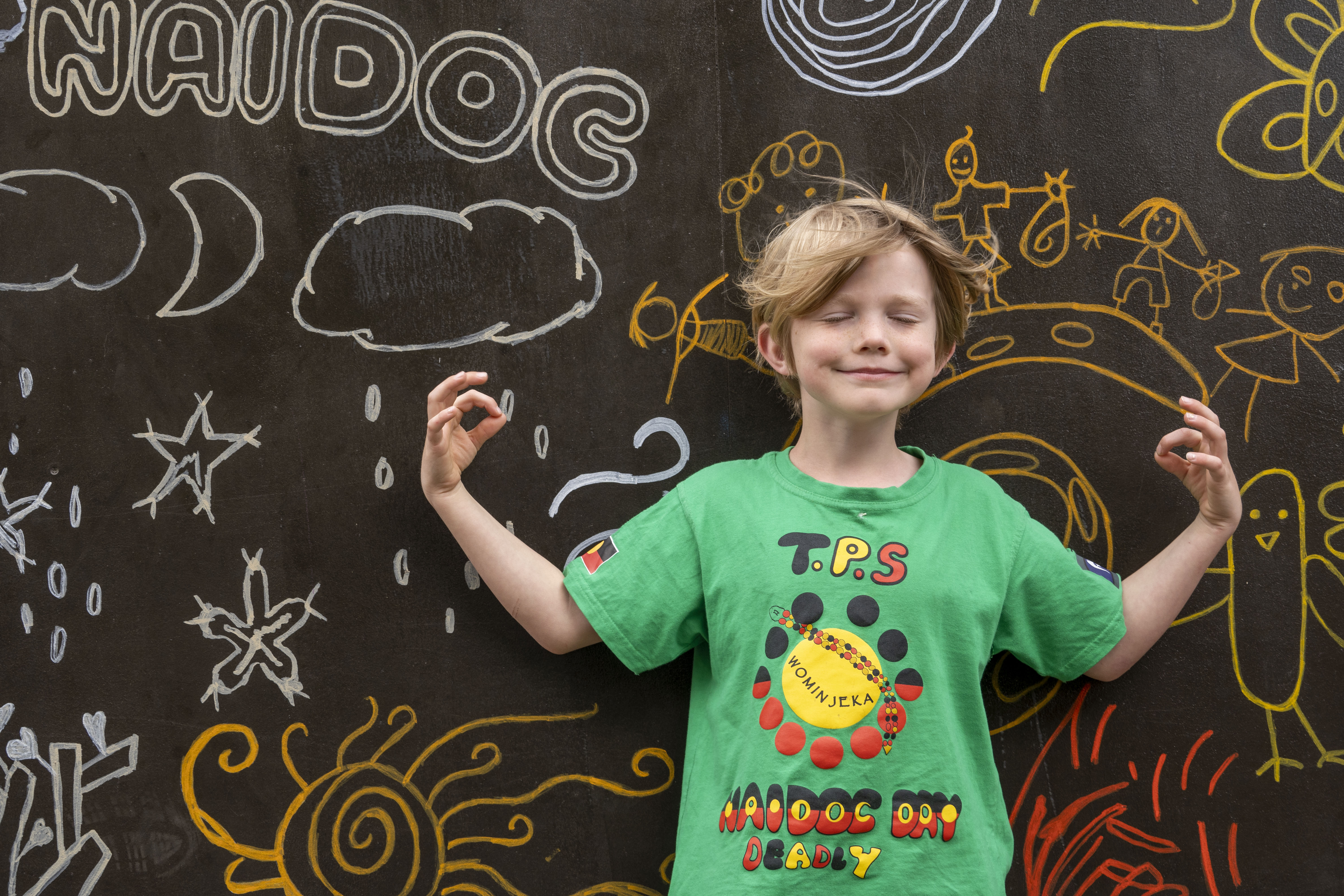 Primary school aged boy in a NAIDOC t-shirt standing in front of a chalk wall covered in children's drawings. He is standing with his eyes closed, a smile on his face, with his arms raised and his index fingers touching his thumbs.