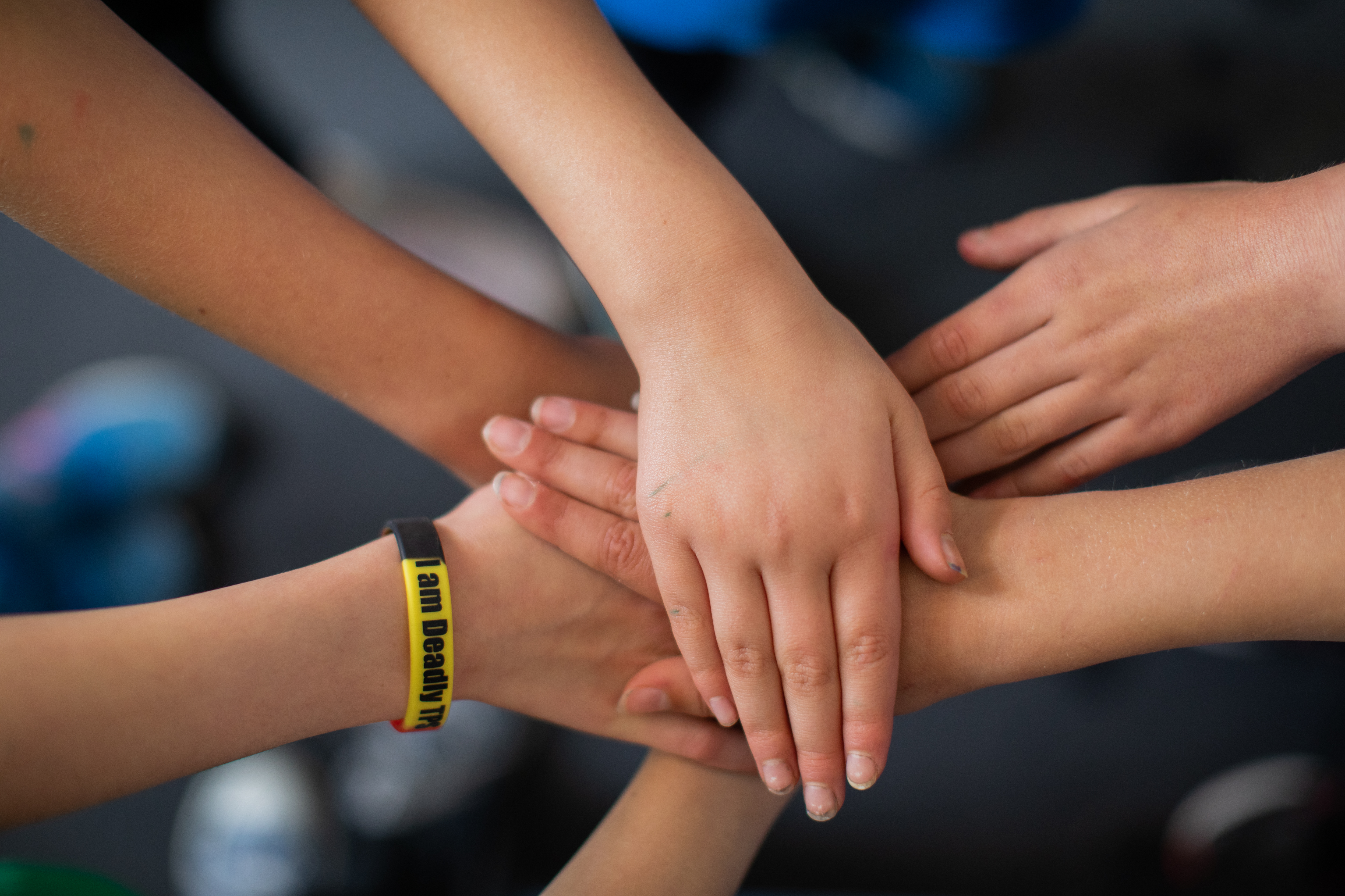 Six hands stacked on top of each other. One child is wearing a wrist band that says 'I am deadly' in black, yellow and red.