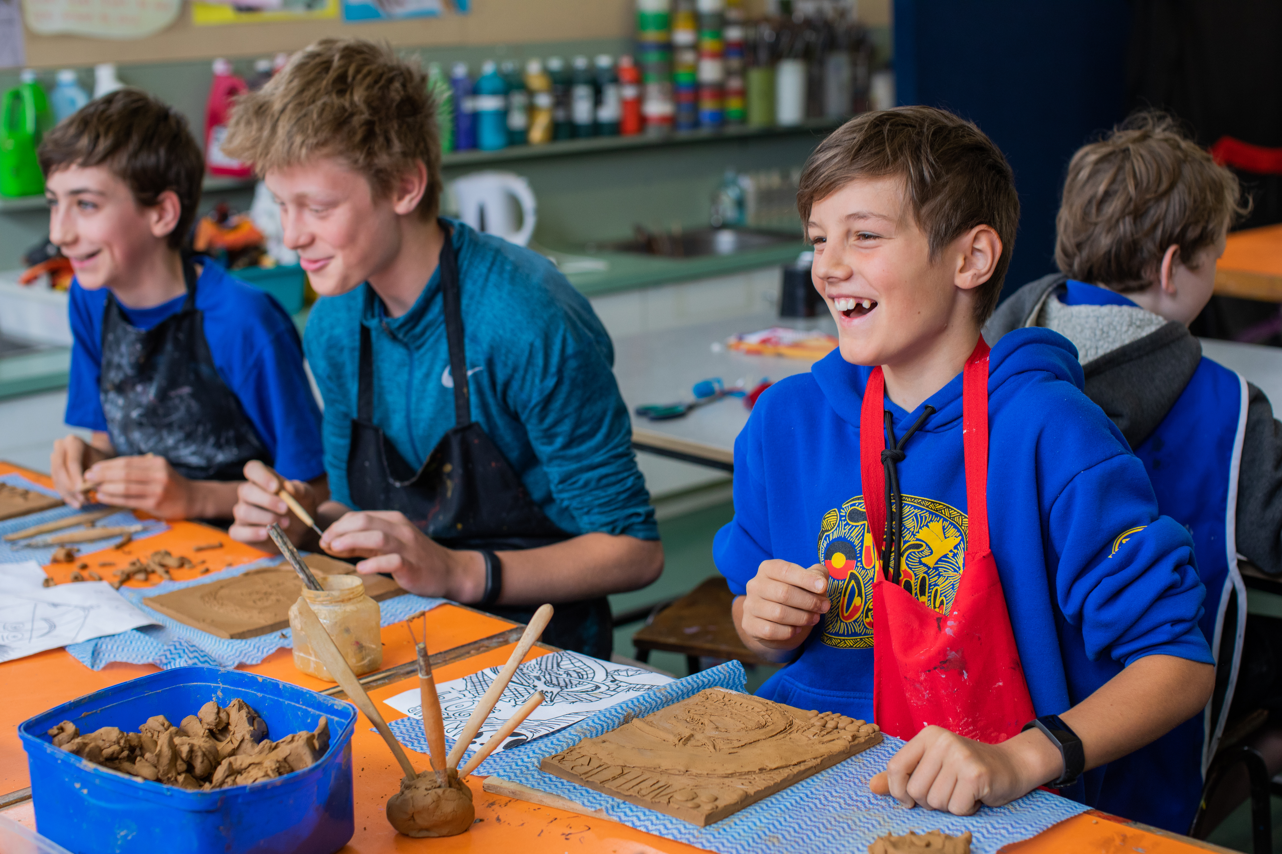 Three teenage boys sitting at a table in an Art classroom wearing aprons and working with clay.