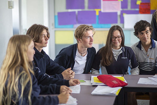 A group of adolescents sitting at desks, speaking with the teacher