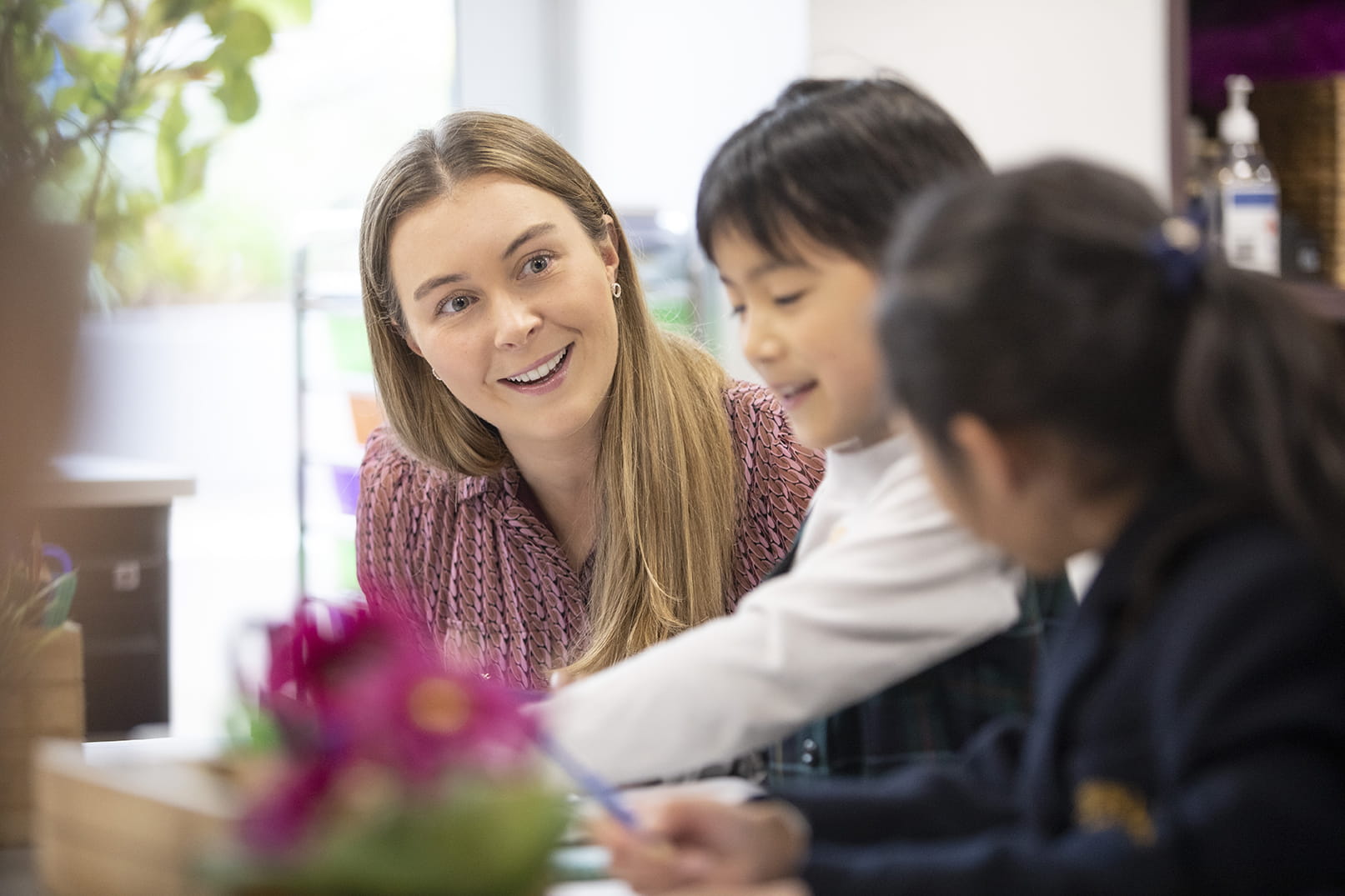 The image shows a woman with long blonde hair smiling and engaging with two young children in a classroom setting. The woman is wearing a patterned pink blouse and appears to be guiding or assisting the children with an activity. One child, a boy, is looking down and working on something, while the other child, a girl, is partially out of focus. The background includes a blurred potted plant and classroom supplies, creating a warm and supportive learning environment.